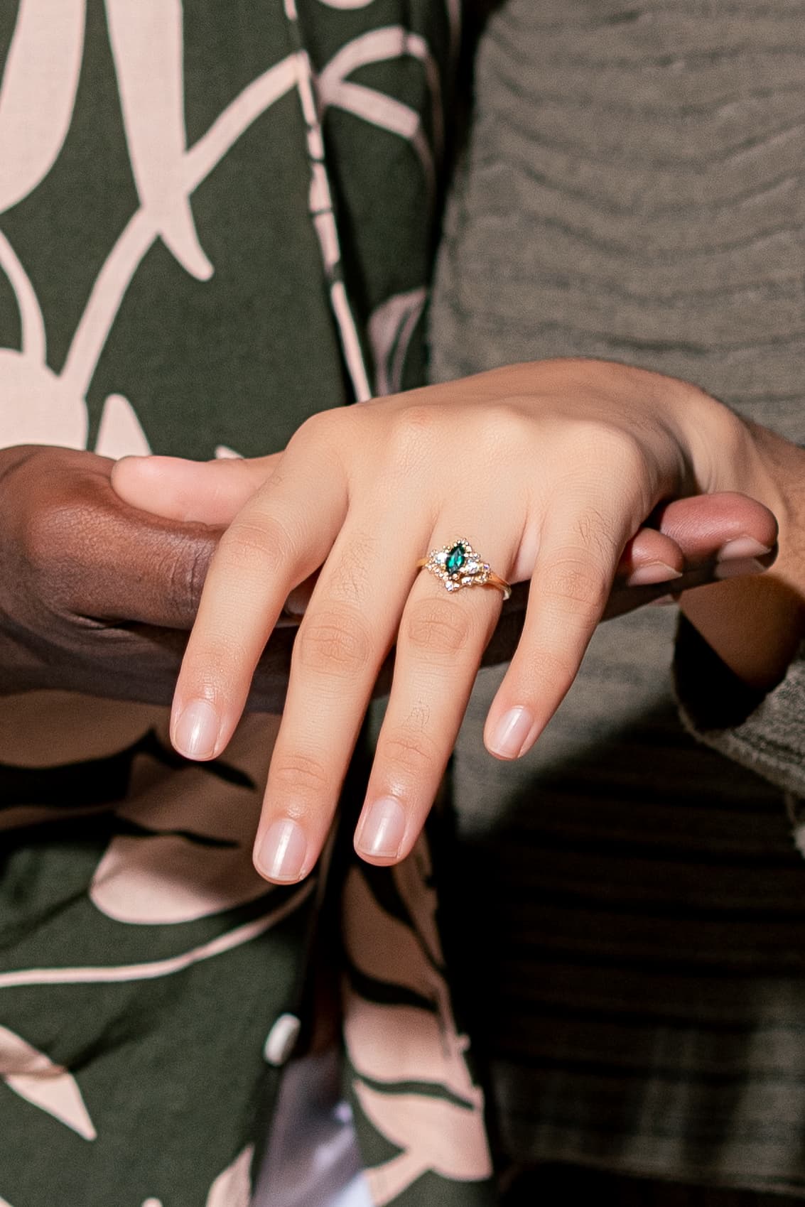 A hand wearing a green gemstone engagement ring rests on top of another person's hand.