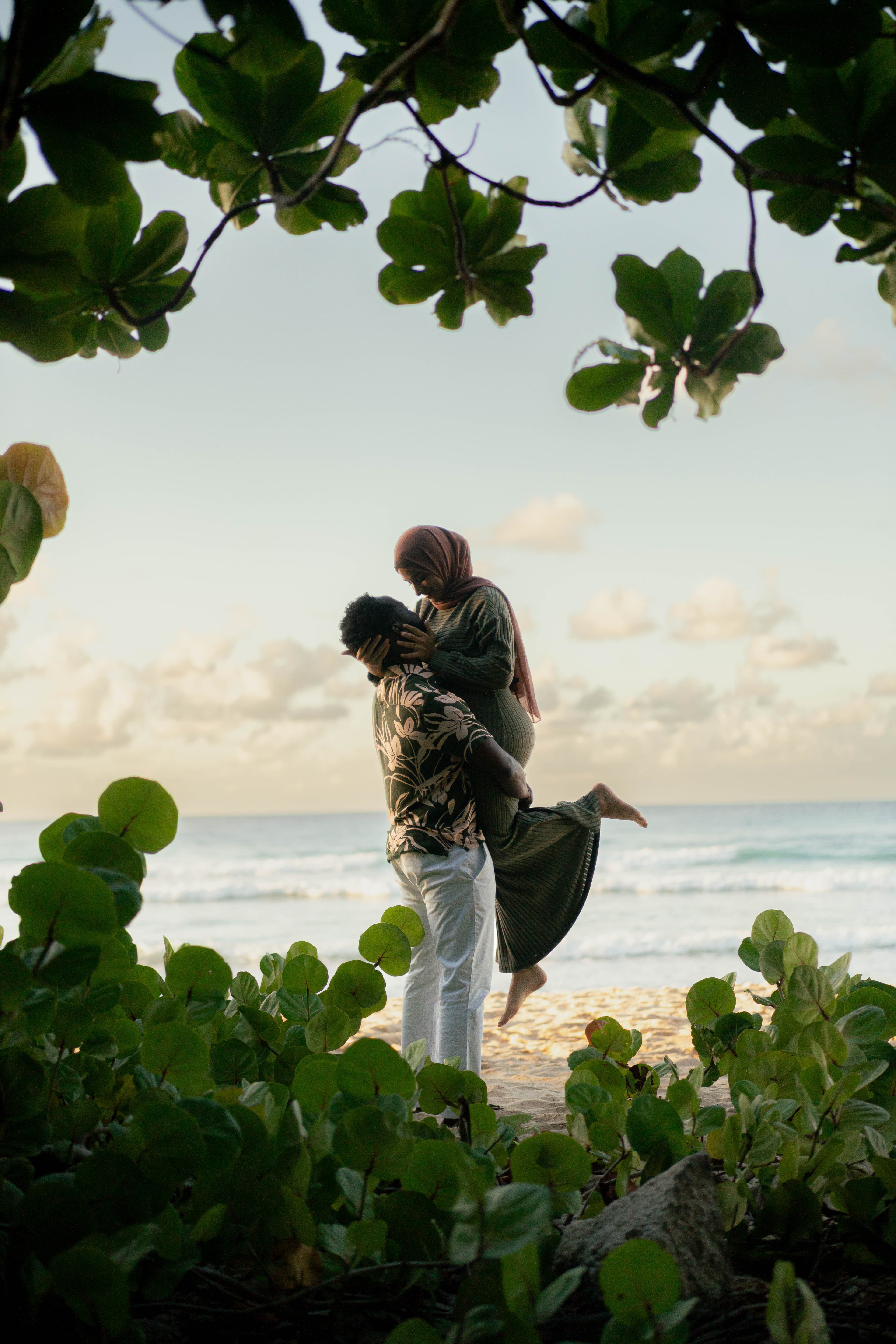 Man lifts a woman wearing a hijab on a beach, framed by lush green leaves.