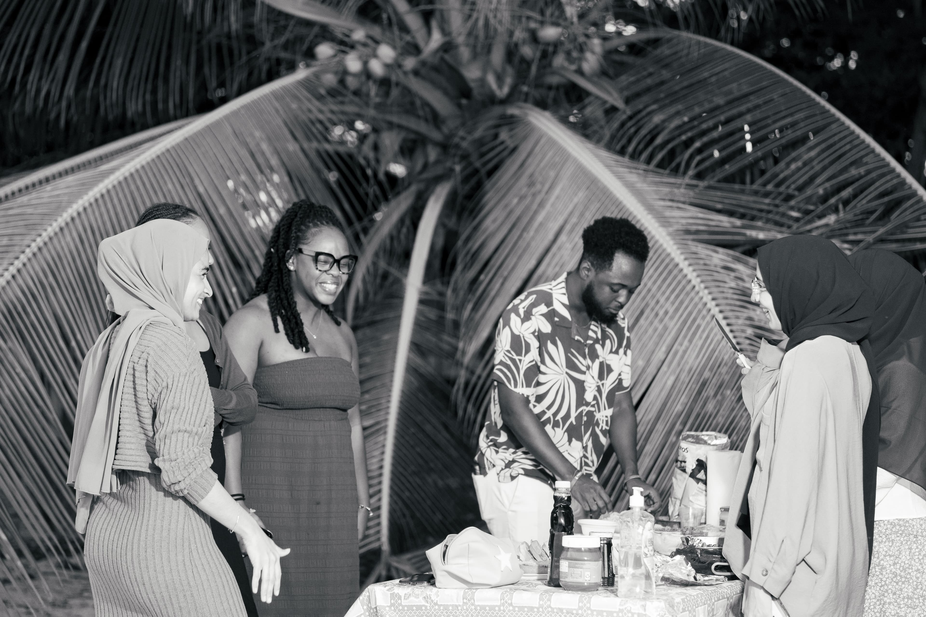 Friends gather around a table outdoors at night, smiling and preparing food under palm fronds.