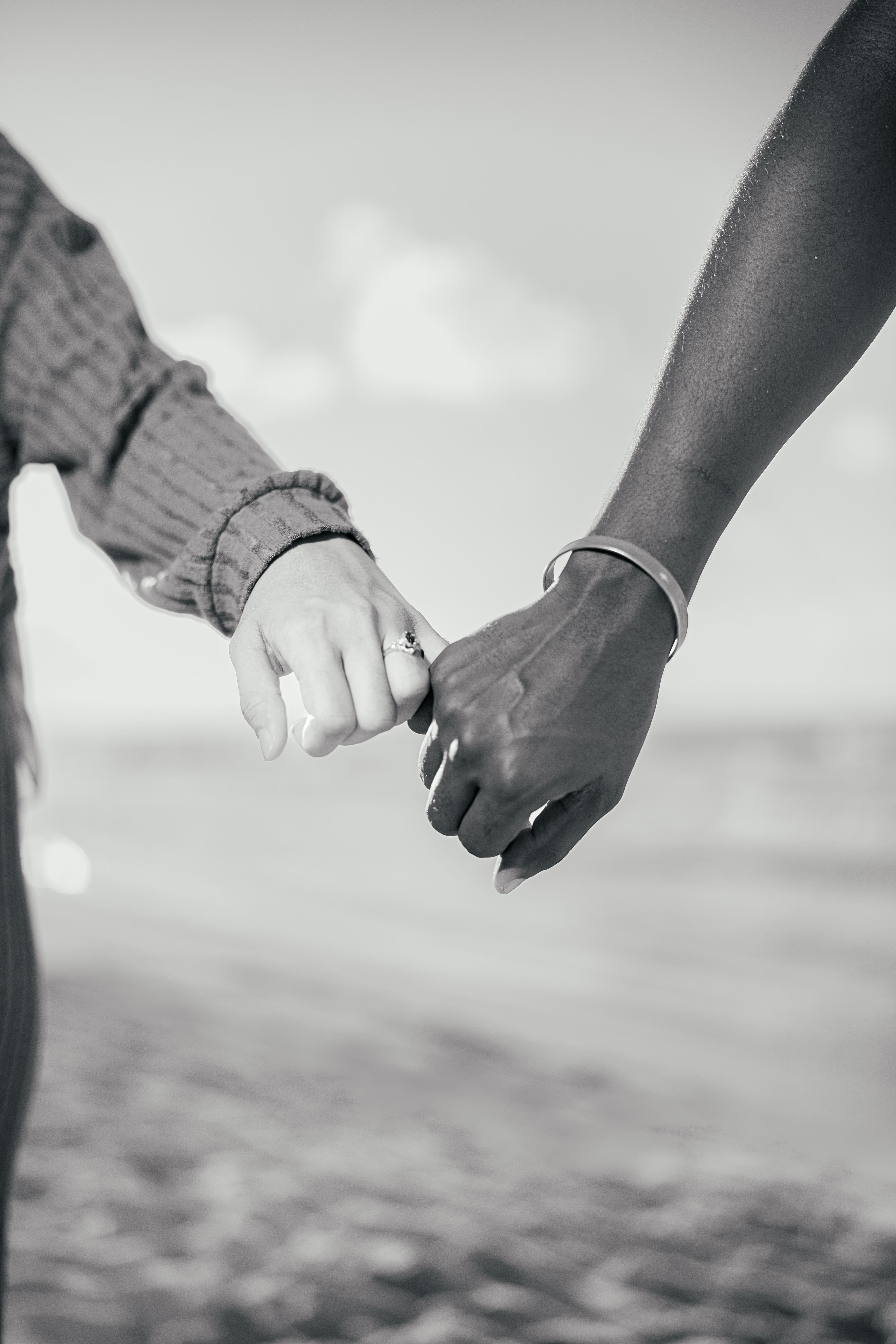 Black and white close-up of two people linking pinky fingers on a blurred beach.