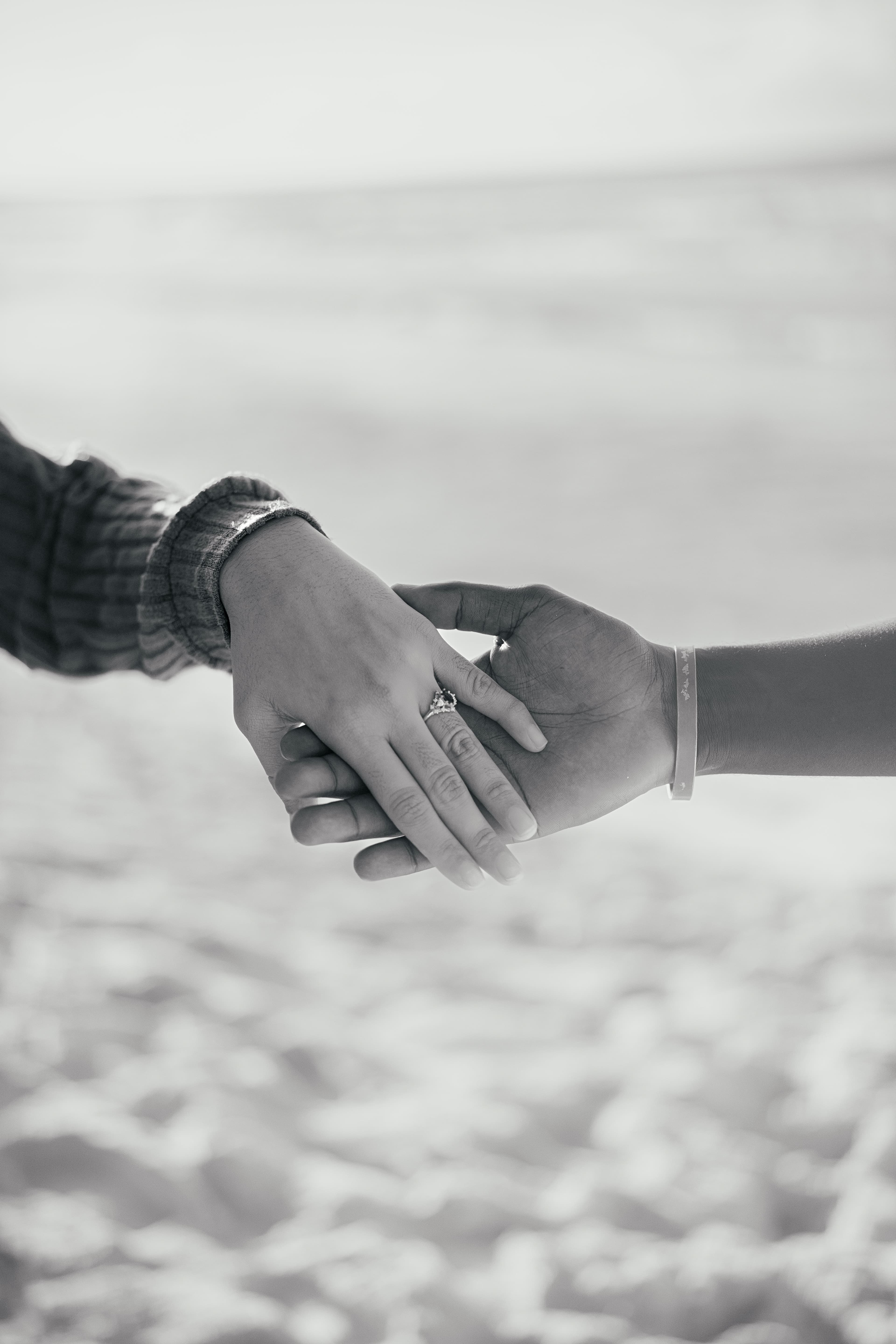 Black and white close-up of two hands holding, featuring an engagement ring on a beach.