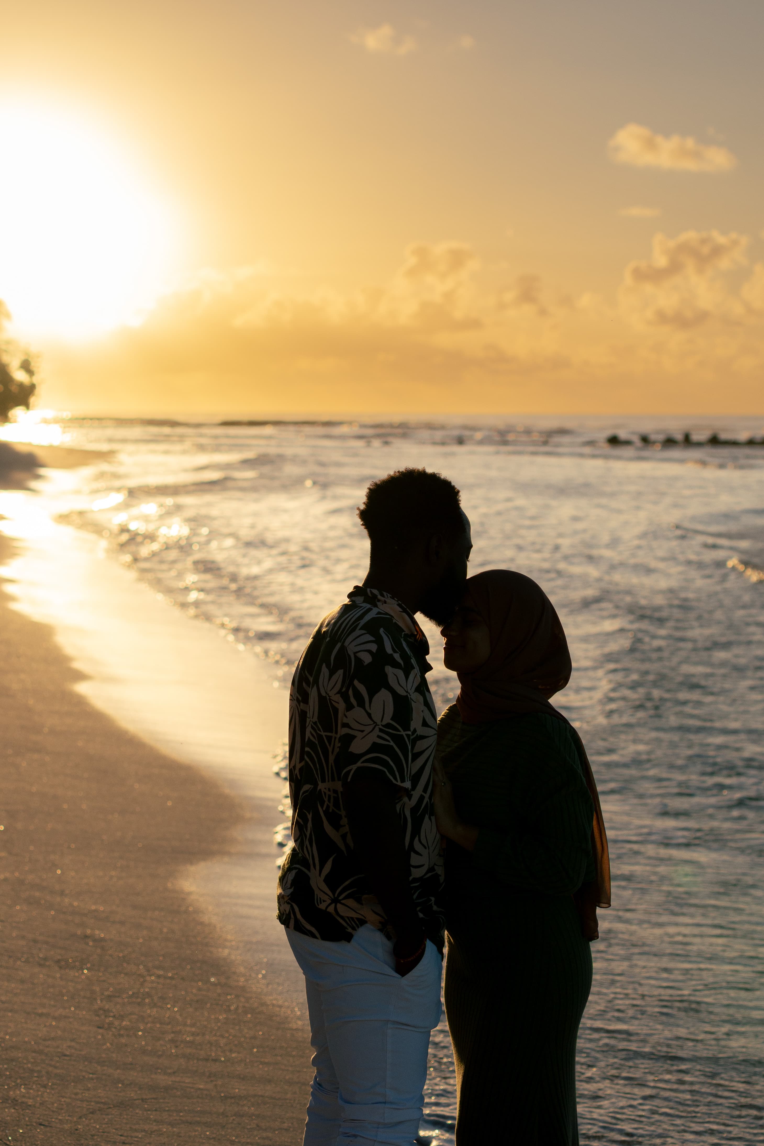 Man kissing woman in hijab on a beach at sunset, silhouetted against the golden sky.